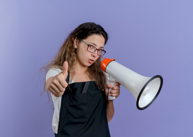 Foto gratuita donna giovane parrucchiere professionista in grembiule che parla al megafono che mostra i pollici in su