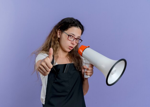 親指を立ててメガホンに話しかけるエプロンの若いプロの美容師の女性