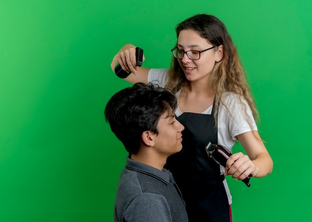 Free photo young professional hairdresser woman in apron sprinkling water on hair of man client using spray before haircut