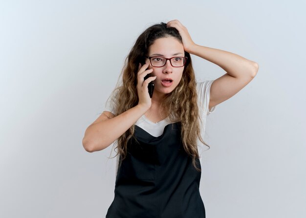 Young professional hairdresser woman in apron talking on mobile phone being confused standing over white wall