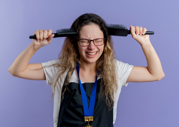 Foto gratuita giovane donna parrucchiere professionista in grembiule con medaglia d'oro intorno al collo che tiene le spazzole per capelli vicino alla testa che sembra delusa