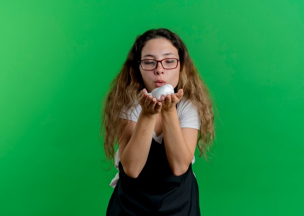 Donna giovane parrucchiere professionista in grembiule con schiuma da barba nelle mani che soffia su di esso in piedi sopra la parete verde