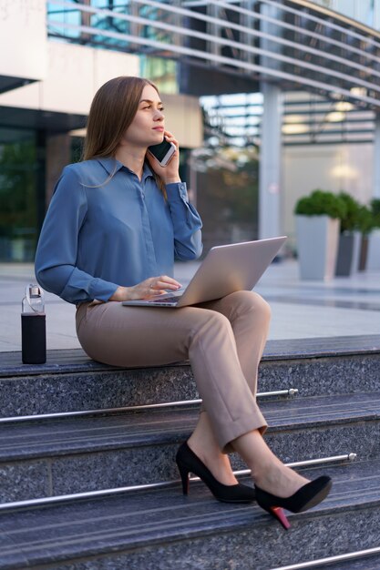 젊은 전문 여성 유리 건물 앞 계단에 앉아 무릎에 노트북을 들고 휴대 전화로 이야기