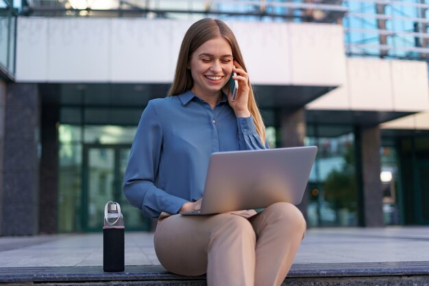 Молодая профессиональная женщина сидит на лестнице перед стеклянным зданием, держит ноутбук на коленях и разговаривает по мобильному телефону