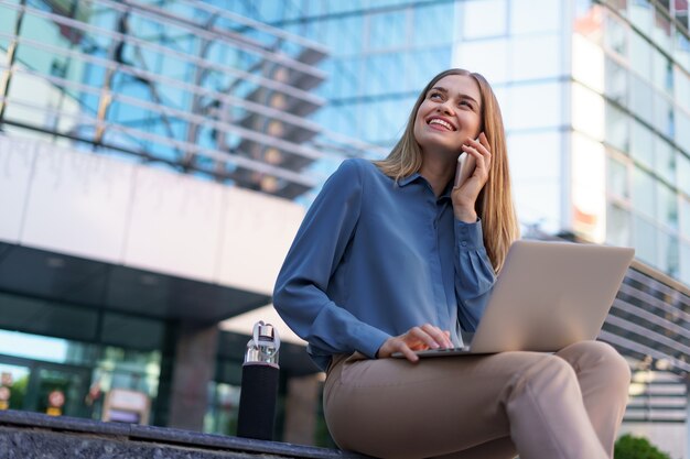 Молодая профессиональная женщина сидит на лестнице перед стеклянным зданием, держит ноутбук на коленях и разговаривает по мобильному телефону