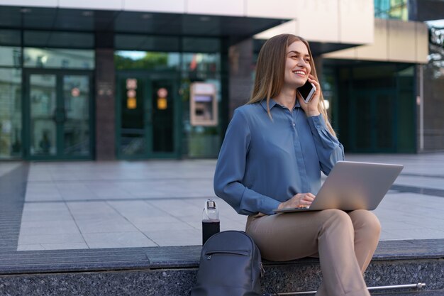 young professional woman sitting on stair in front of glass building, holding laptop in lap and talking on mobile phone