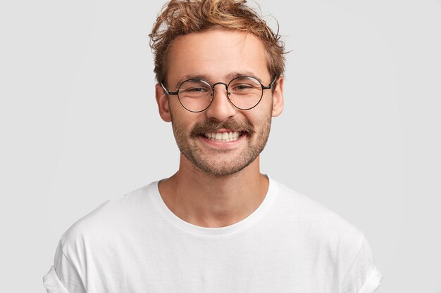 Young prosperous businessman with curly hair, bright smile, glad to achieve success in life, dressed in white t shirt, enjoys day off at home with family