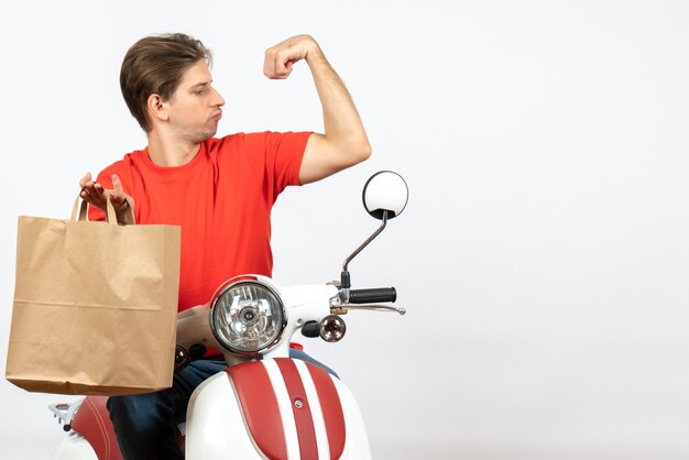 Young proud delivery guy in red uniform sitting on scooter holding paper bag and showing his muscular on white wall