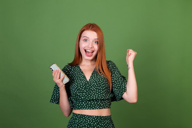 興奮した携帯電話のテキストメッセージチャットで緑の壁に若い赤い髪の女性は勝者のジェスチャーを示しています