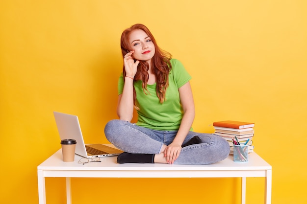 Young red haired female wearing green t shirt and jeans, satisfied student being tired from learning for long time