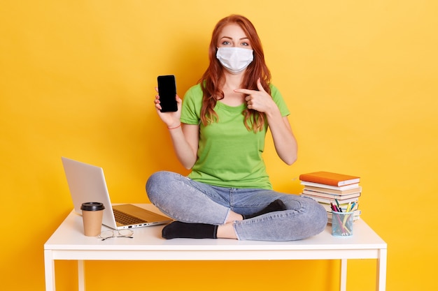 Young red haired woman studying while sitting on white table, holding phone with blank screen, surrounded books, lap top, wearing medical mask isolated over yellow background.