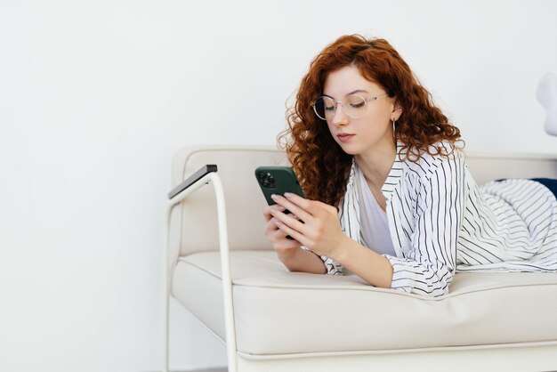 Young red haired woman using smart phone while resting on sofa at home