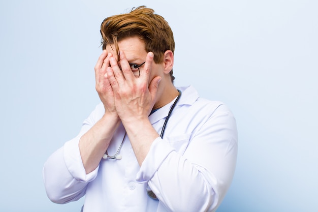 Premium Photo | Young red head doctor feeling scared or embarrassed,  peeking or spying with eyes half-covered with hands on blue wall