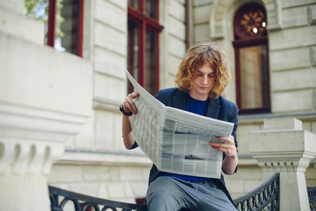 Young reddish man reading newspaper near old style building