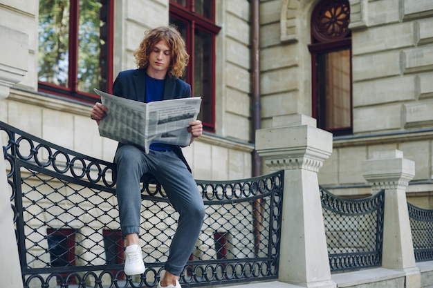 Young reddish, red haired attractive man with curly hair, reading a newspaper sitting near an urban old style building. Youth in action young leader getting to know world, city news.