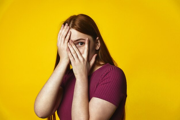 young redhead caucasian girl is covering face with hands and looking through the fingers