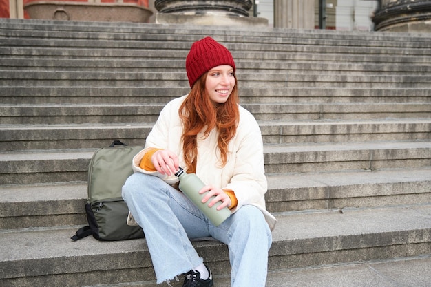 Young redhead female tourist rests during her trip opens thermos and drinks hot tea having a break a