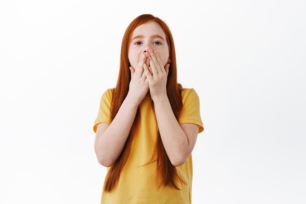 Молодая рыжая девушка прикрывает рот и задыхается, потрясенно смотрит в камеру, удивляется и поражается, стоя в желтой футболке на белом фоне