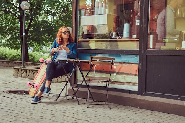 若い赤毛の女の子は、コーヒーショップの近くに座って、スケートボードに乗った後にリラックスして、コーヒーを飲みます。