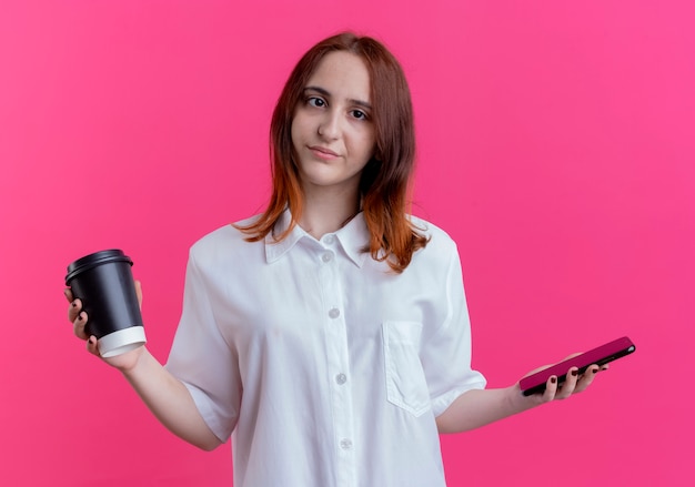 young redhead girl holding phone and cup of coffee isolated on pink wall