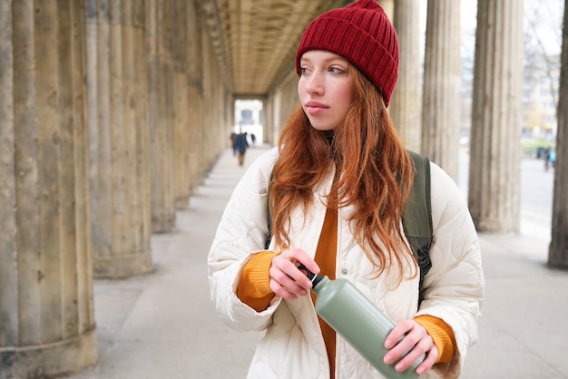Free photo young redhead girl holds thermos in hands pours herself a hot drink while walking in city tourist ta