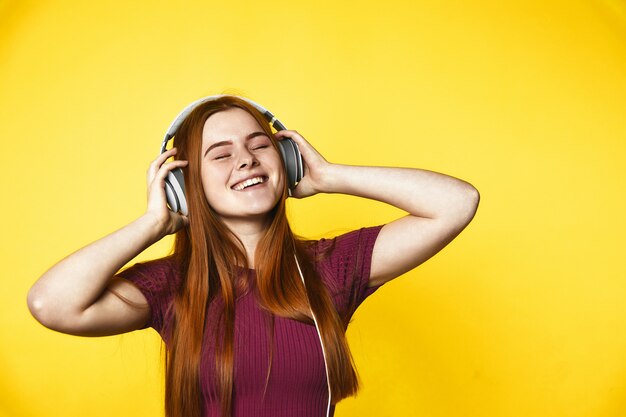 젊은 빨간 머리 소녀는 닫힌 눈을 가진 헤드폰을 통해 행복하고 듣는 음악입니다