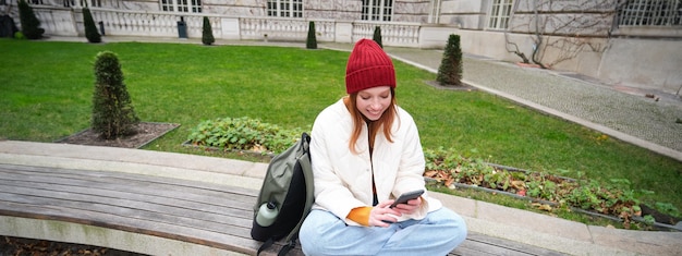 無料写真 若い赤毛の女子学生がベンチに座り、スマートフォンのアプリを使用してオンラインでビデオを視聴し、メッセージを送信