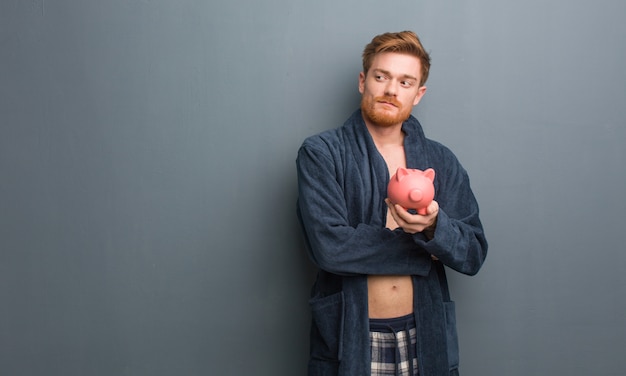 Young redhead man wearing pajama smiling confident and crossing arms, looking up. he is holding a piggy bank.