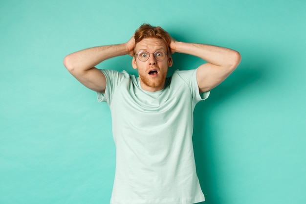 Young redhead man with beard, wearing glasses and t-shirt, holding hands on head and staring in panic, standing alarmed and anxious against turquoise background.