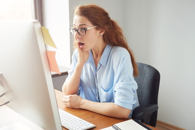 Young redhead office worker wearing glasses and blue shirt, sitting with surprised expression