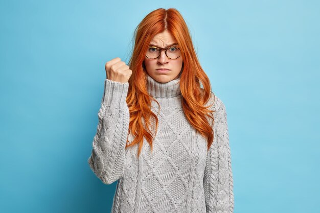 젊은 빨간 머리 여자는 분노로 주먹을 쥐고 캐주얼 니트 스웨터를 입은 불쾌한 표정으로 보입니다.