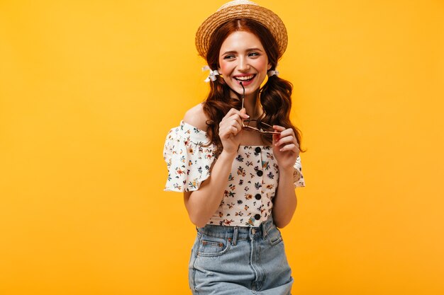 Young redhead woman in great mood smiles and holds glasses. Lady in straw hat and floral print shirt looking away.