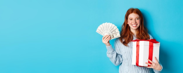 Free photo young redhead woman holding christmas gift box and money smiling pleased standing over blue backgrou