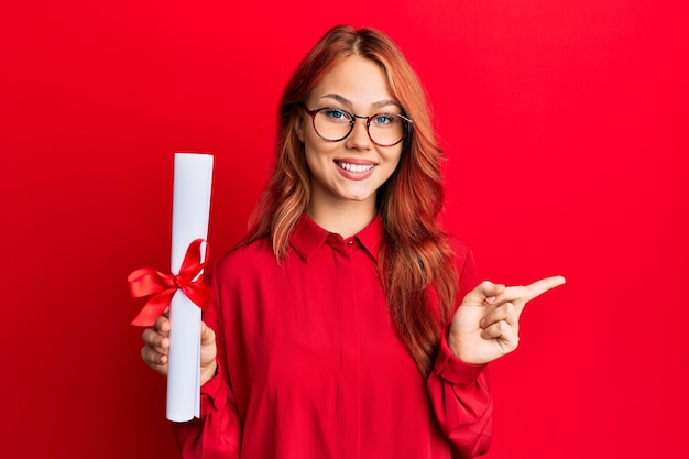 Foto gratuita giovane donna rossa che tiene il diploma di laurea laureato sorridente felice indicando con la mano e il dito di lato