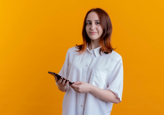 Young redhead woman holding phone isolated on yellow