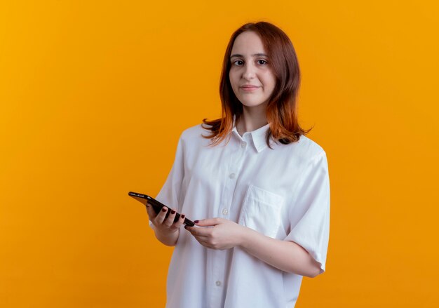 young redhead woman holding phone isolated on yellow