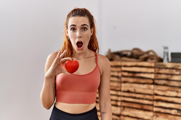 Free photo young redhead woman holding red heart at the gym scared and amazed with open mouth for surprise disbelief face