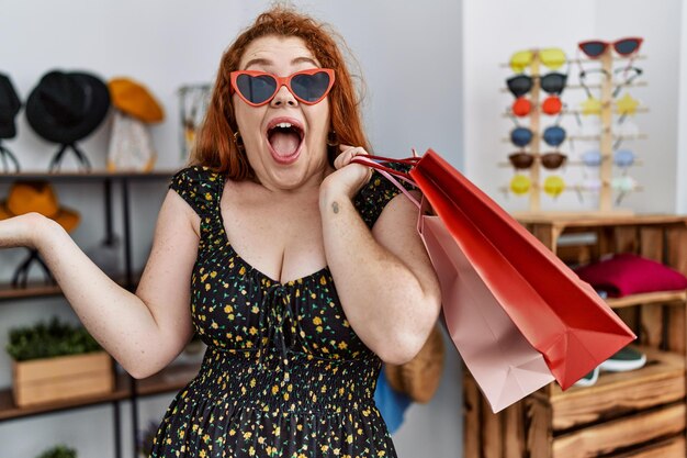 소매점에서 쇼핑백을 들고 있는 젊은 빨간머리 여성은 행복한 미소와 손을 들어 승자 표현으로 성취를 축하합니다.