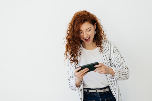 Giovane donna rossa che gioca ai videogiochi sul suo telefono guardando il video in diretta streaming sul cellulare ridendo e sorridendo guardando lo sfondo bianco dello schermo mobile