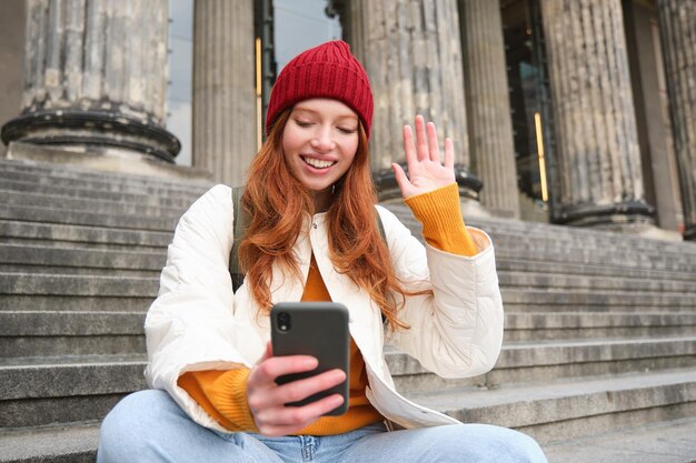 若い赤毛の女性は屋外の階段に座って、スマートフォンのカメラのビデオチャットで手を振っています