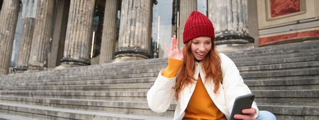Foto gratuita una giovane donna dai capelli rossi si siede sulle scale all'aperto e agita la mano davanti alle chat video della fotocamera dello smartphone