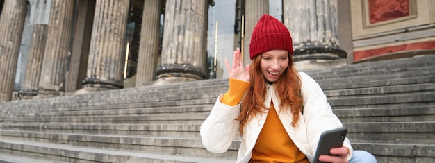 Free photo young redhead woman sits on stairs outdoors and waves hand at smartphone camera video chats with