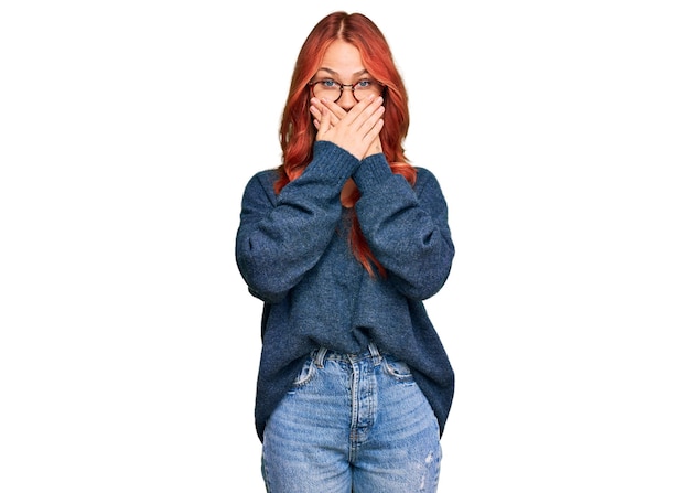 Молодая рыжая женщина в повседневном свитере и очках потрясена, прикрывая рот руками по ошибке. секретная концепция.