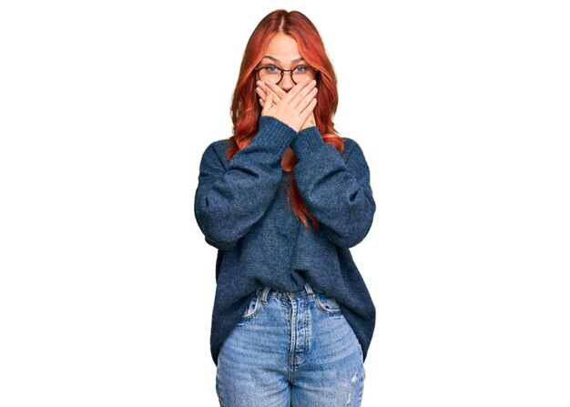 Young redhead woman wearing casual sweater and glasses shocked covering mouth with hands for mistake. secret concept.