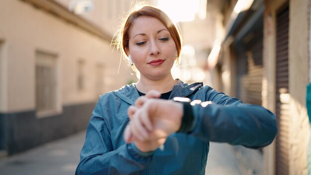 Young redhead woman wearing sportswear listening to music looking stopwatch at street
