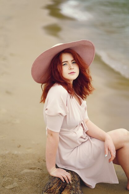 Молодая рыжеволосая девушка в большой круглой шляпе и розовом платье, сидящем на пляже у океана