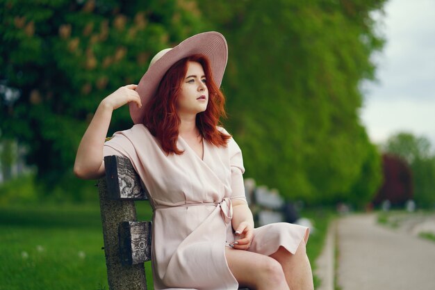 Молодая рыжеволосая девушка в большой круглой шляпе и розовом платье, сидящем на скамейке