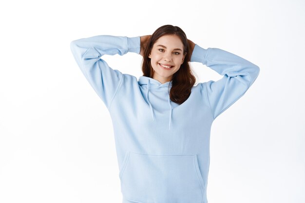 Young relaxed girl student having leisure time, holding hands behind head as if lying and resting, smiling happy at front, standing against white wall