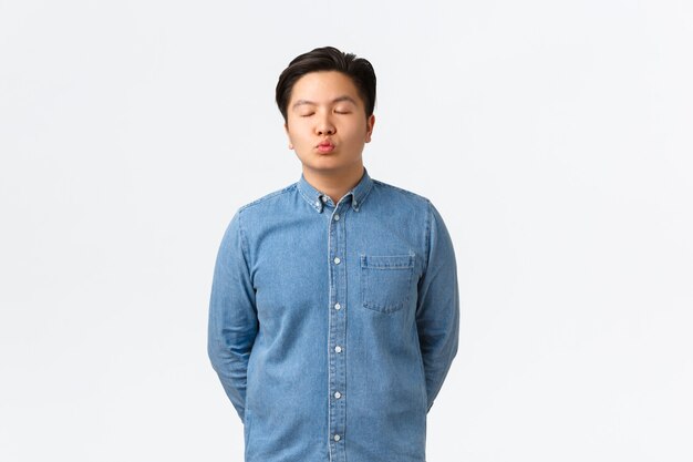 Young romantic asian guy in blue shirt, close eyes and pouting as waiting for kiss, having date with girlfriend, confessing feelings, leaning towards camera mwah, standing white background.
