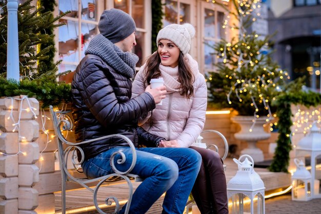 Молодая романтическая пара в теплой одежде сидит на скамейке на вечерней улице, украшенной красивыми огнями, разговаривает и согревается кофе на Рождество на открытом воздухе